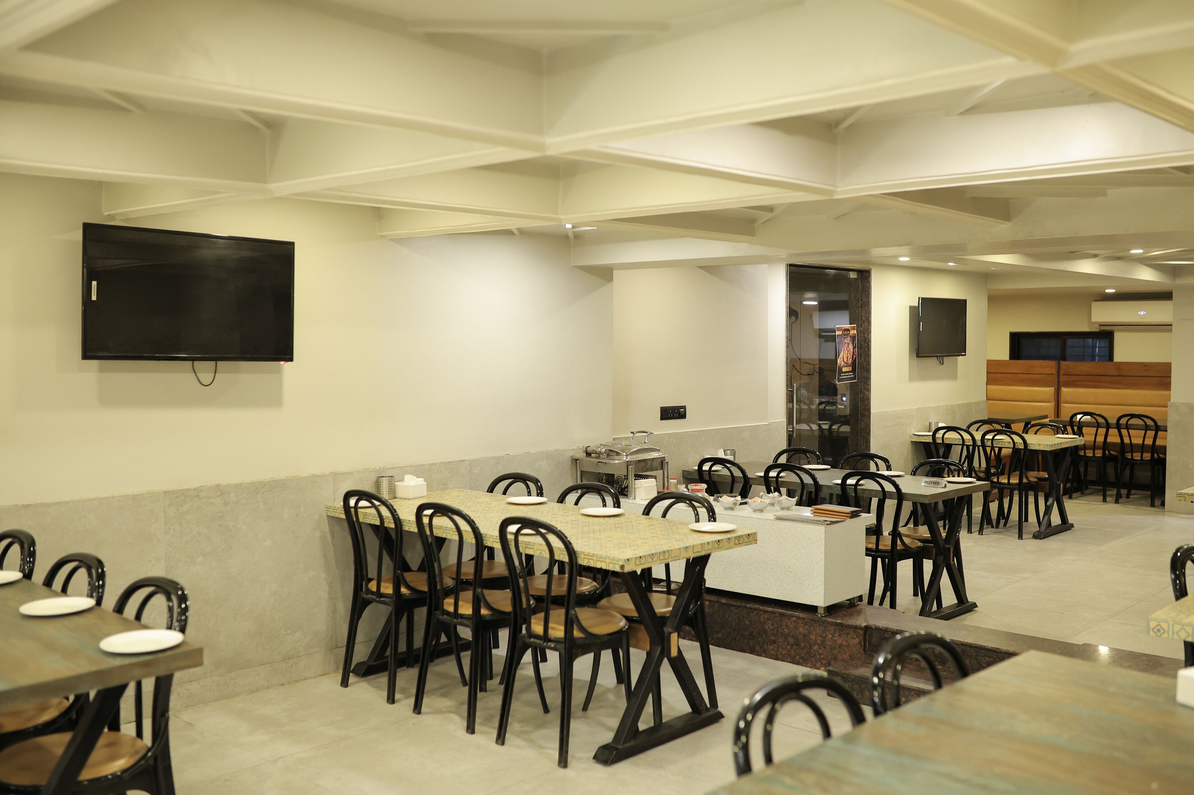 A dining area with multiple tables and chairs, a kitchen area, and a television mounted on the wall.