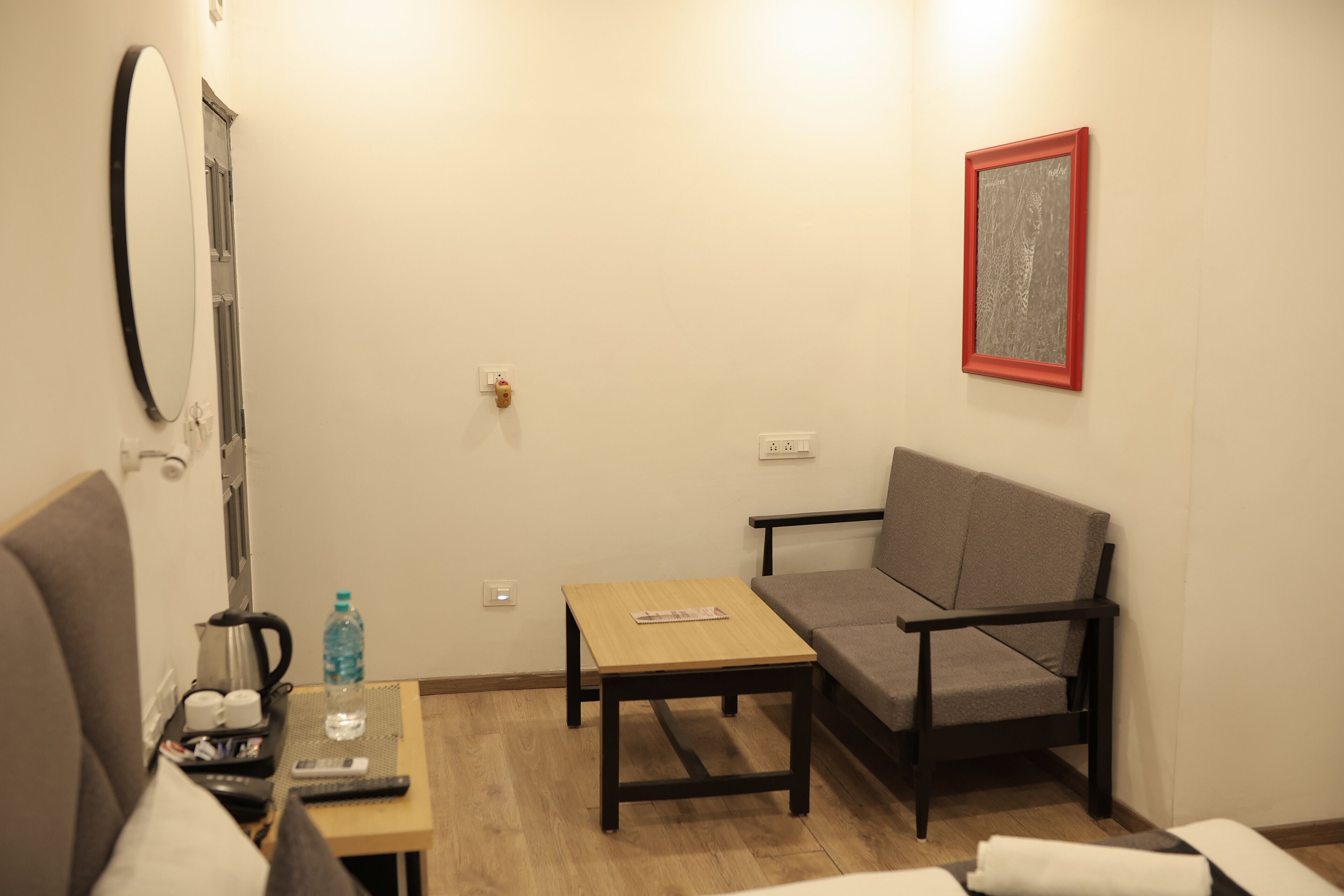 A small, furnished room with a sofa, a table with a remote control, a kettle, and a water bottle.