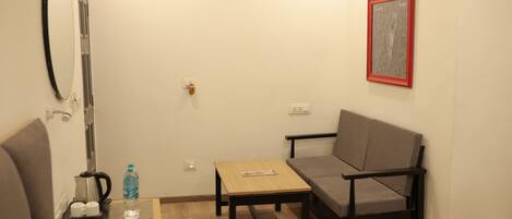 A small, furnished room with a sofa, a table with a remote control, a kettle, and a water bottle.