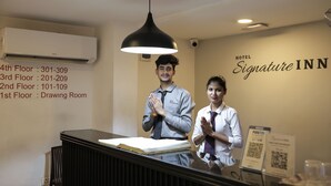 Un comptoir d’accueil d’hôtel avec un luminaire suspendu noir, un climatiseur fixé au mur et une enseigne indiquant le nom de l’hôtel : « Signature INN ».