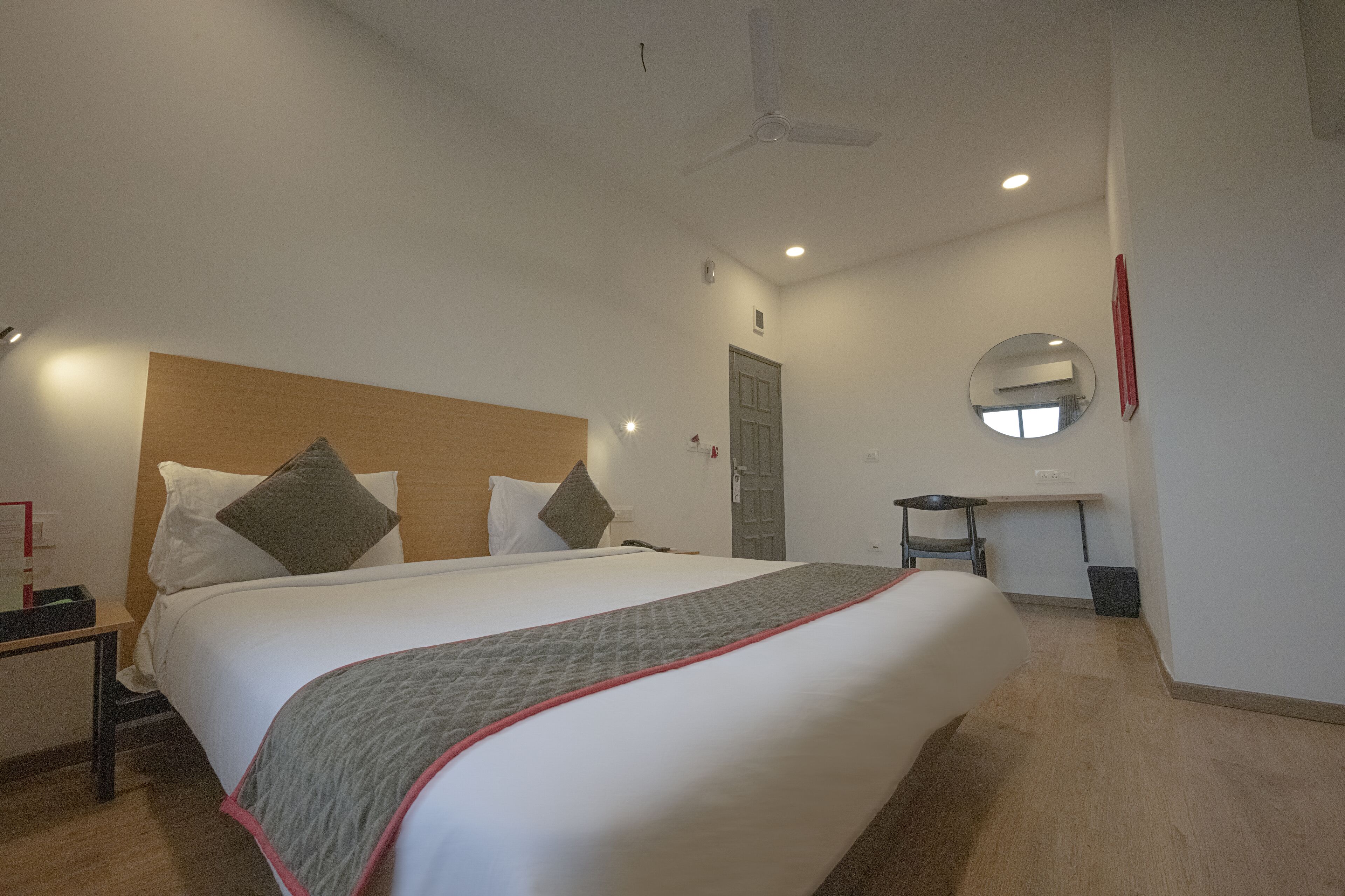 A hotel room with a large bed, a bedside table, a desk, a mirror, and a ceiling fan.