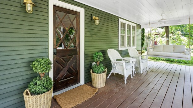 A porch with a green exterior, a dark wooden door, wicker baskets with plants, white wicker chairs, and a white sofa. - Pro Designer Cottage, 4/10 Mile to New Circle Beach (Lakeside)