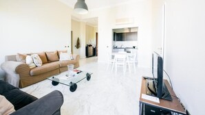 Ein modernes Wohnzimmer mit einer Couch, einem Couchtisch und einem Fernseher. Im Hintergrund ist eine Küche zu sehen.
