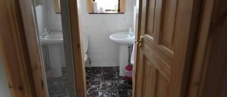 A narrow hallway with wooden doors and a marble floor leading to a bathroom with a toilet, sink, and a window above the sink.