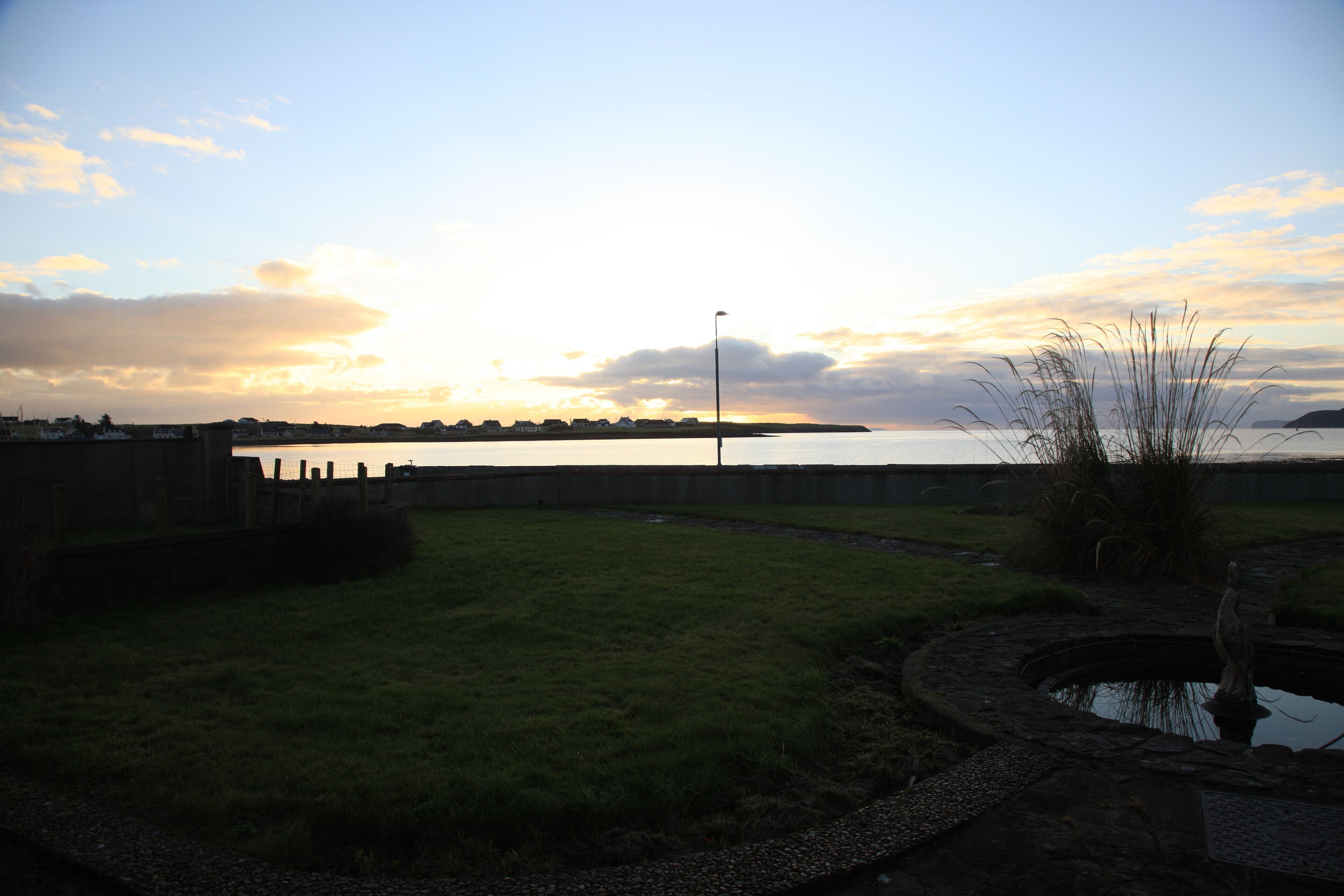 A grassy area with a pond, a statue, and a distant view of a body of water under a cloudy sky.