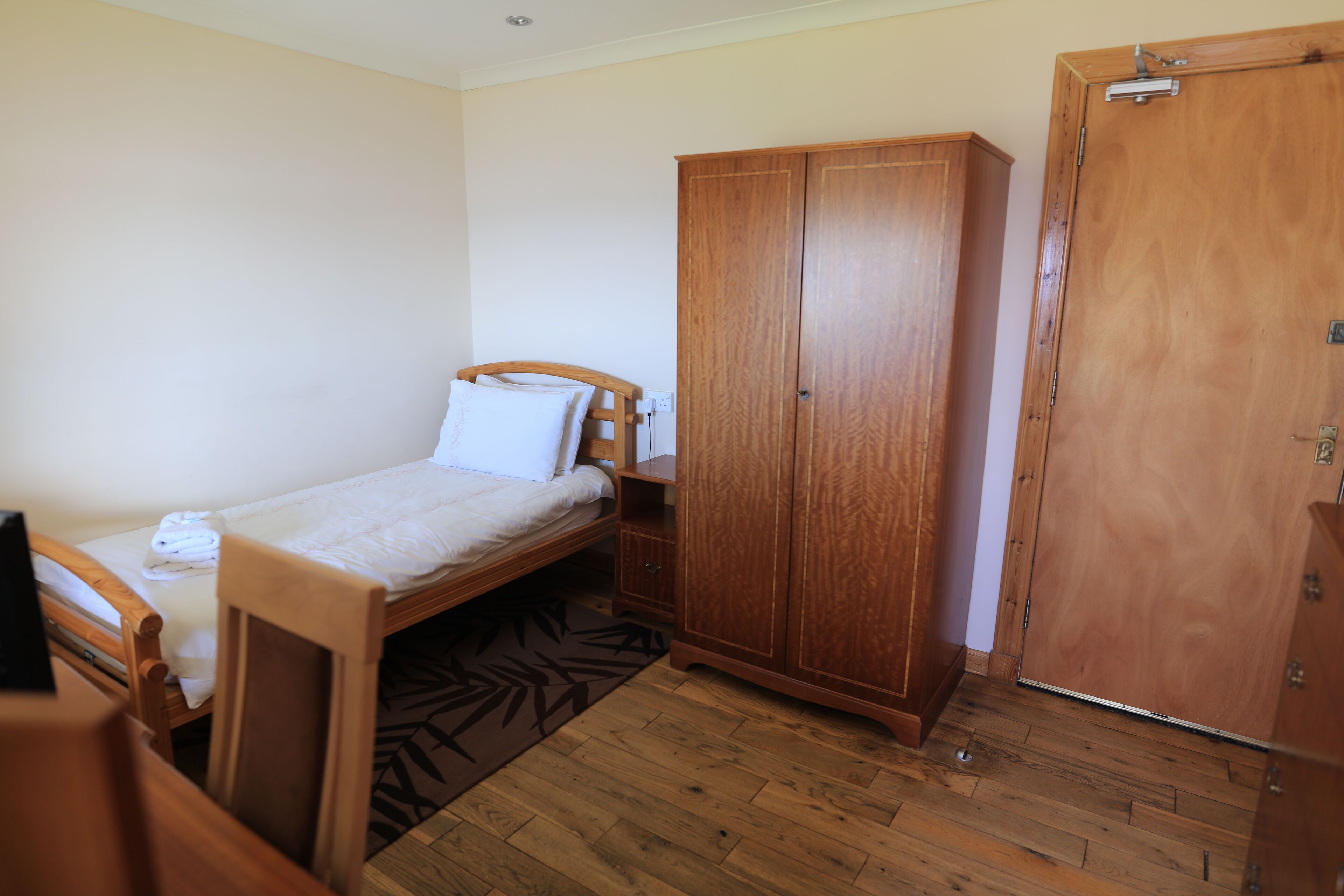 A small, neatly arranged bedroom with a single bed, a wooden wardrobe, and a desk with a chair.