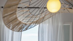 Habitació amb cortines blanques translúcides, una làmpada penjant i vista al mar i edificis.