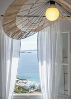 Una habitación con cortinas blancas traslúcidas, una lámpara colgante y vistas al mar y a edificios.
