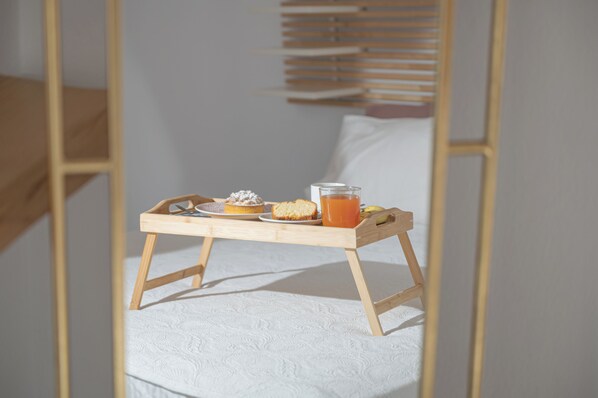 A wooden tray with a slice of cake, a glass of orange juice, and a cookie on a white textured bedspread.