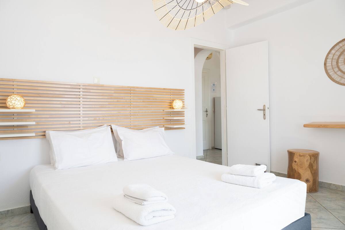Quarto de hotel moderno com uma cama grande, cabeceira de madeira e um pequeno banquinho de madeira.