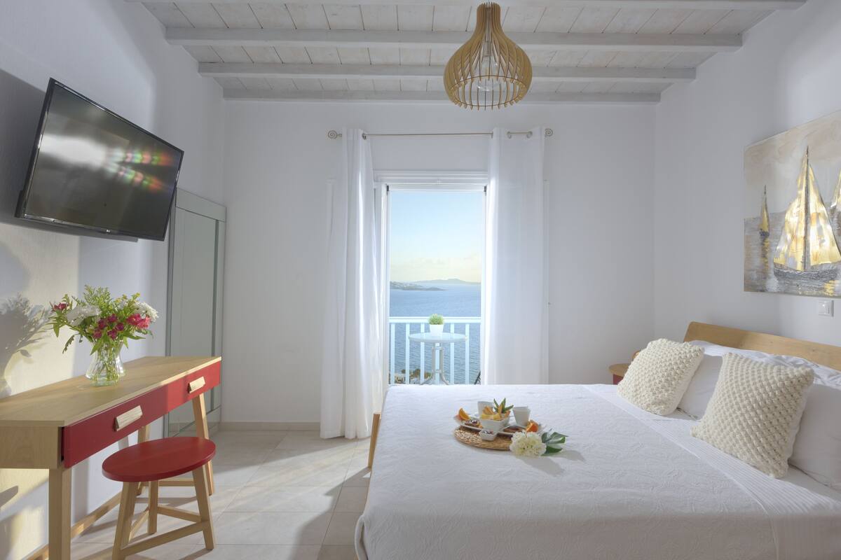 Quarto com cama, escrivaninha, cadeira, televisão e varanda com vista para o mar.