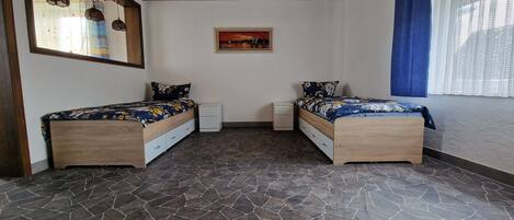 Un dormitorio con dos camas de madera, piso de baldosas, un cuadro en la pared y una ventana con cortinas azules.