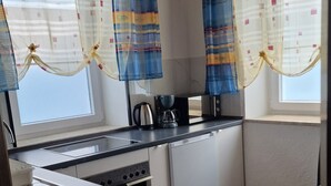 A compact kitchen with a stove, sink, and coffee maker. There are two windows with patterned curtains. The floor has a stone tile pattern. - W.I.L.A. 3 (Helmbrechts)
