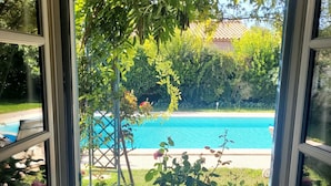 Une vue à travers une fenêtre sur une piscine entourée d’une végétation luxuriante et donnant sur un espace patio.