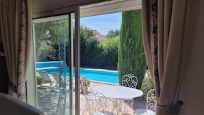 Une pièce dotée d’une porte-fenêtre coulissante donnant sur une terrasse extérieure avec une piscine, des chaises en métal blanc et une table ronde.