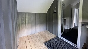 Une petite pièce étroite, dotée d’un plancher en bois et d’un paillasson noir à l’entrée. Une porte donne sur une salle de bain équipée de toilettes et d’un lavabo. Les murs sont peints dans une couleur gris foncé.