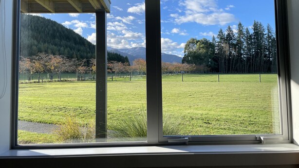 Blick aus dem Fenster auf ein Wiesenfeld mit Bäumen und Bergen im Hintergrund.