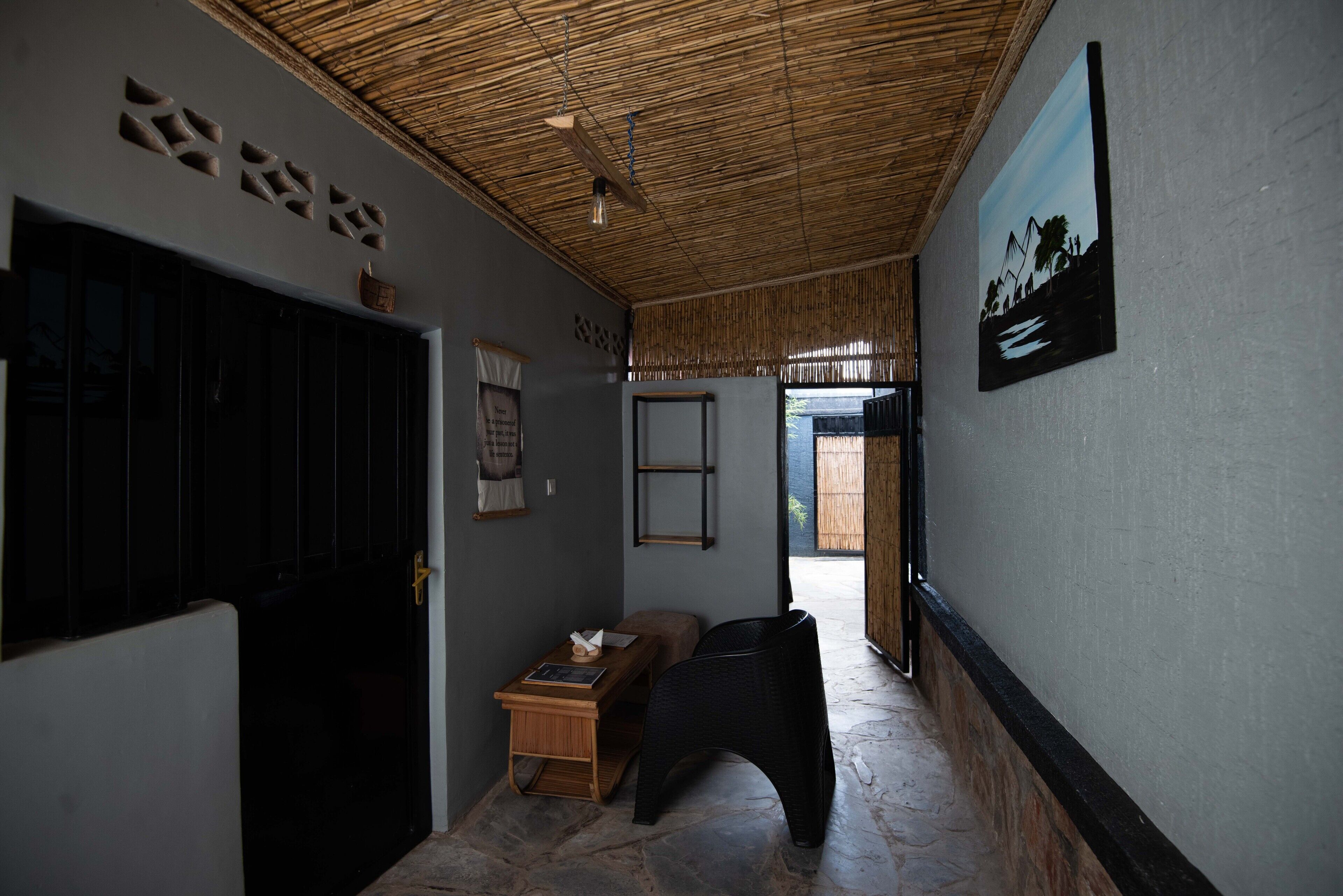 A hallway with a bamboo ceiling, a black chair, a wooden table with a lamp, and a painting on the wall.
