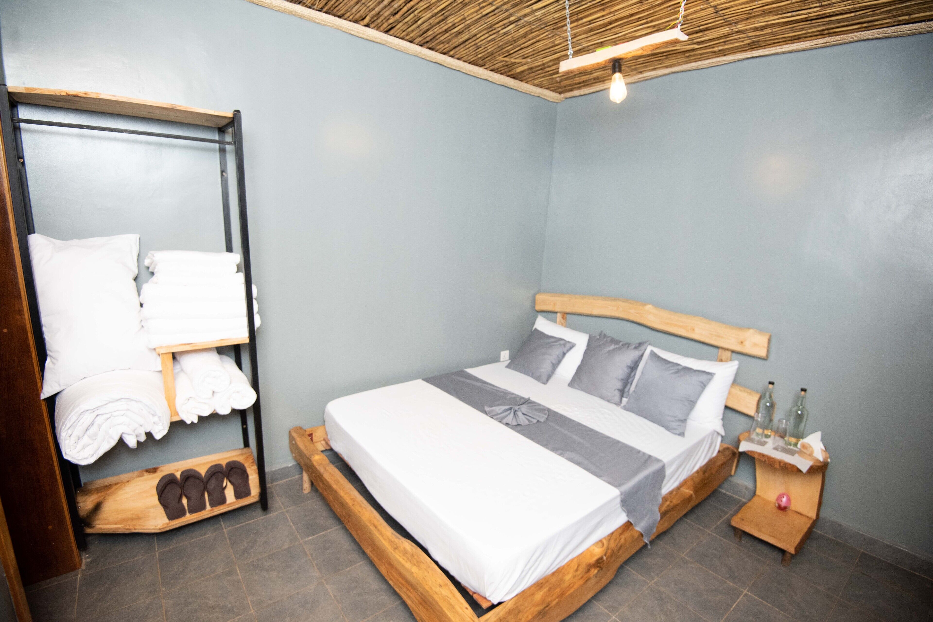 Une chambre à coucher avec un lit en bois, une étagère où sont pliées des serviettes et une petite table de chevet avec des bouteilles.