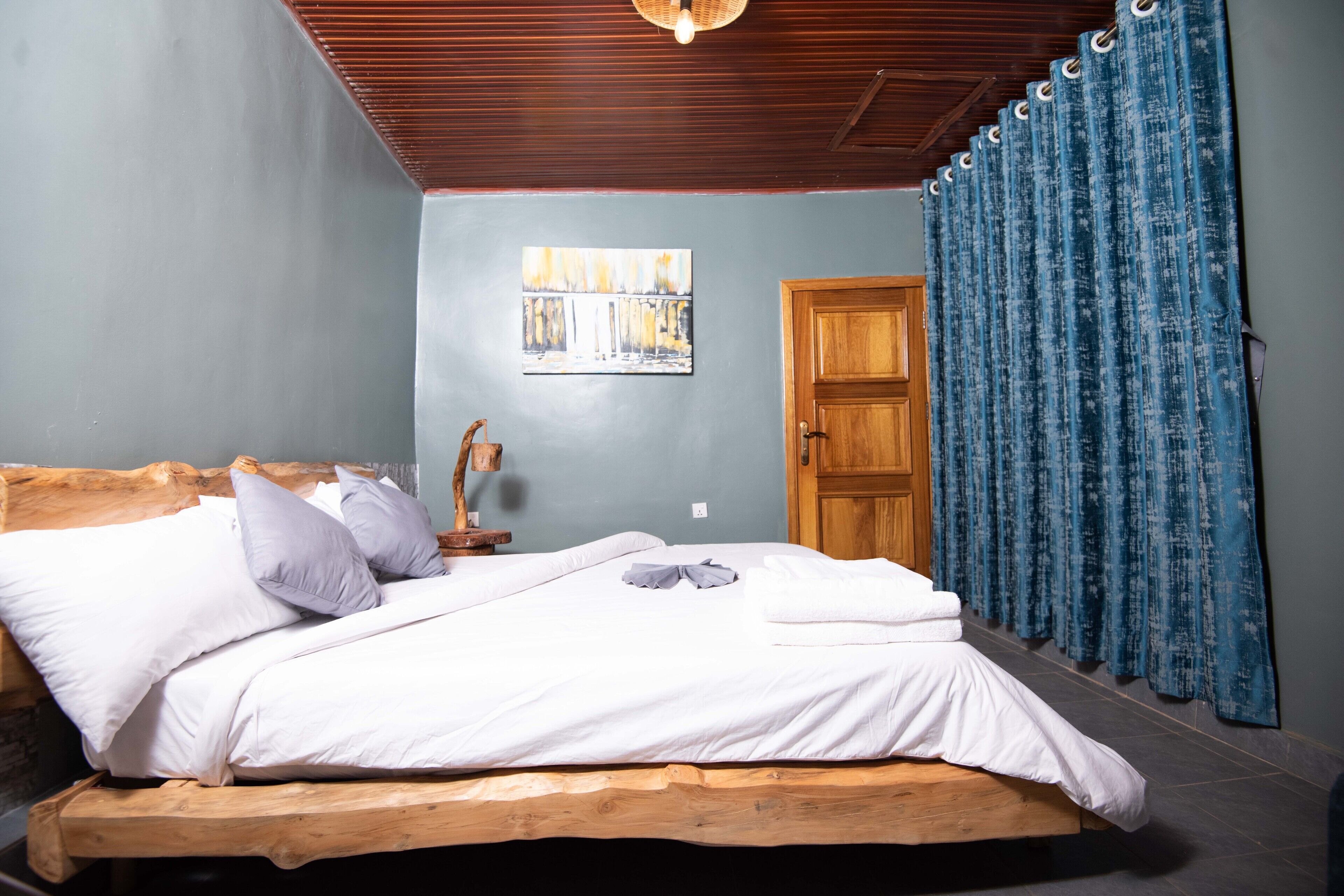 Un dormitorio con estructura de cama de madera, ropa de cama blanca, cabecera de madera, puerta de madera y cortinas azules.