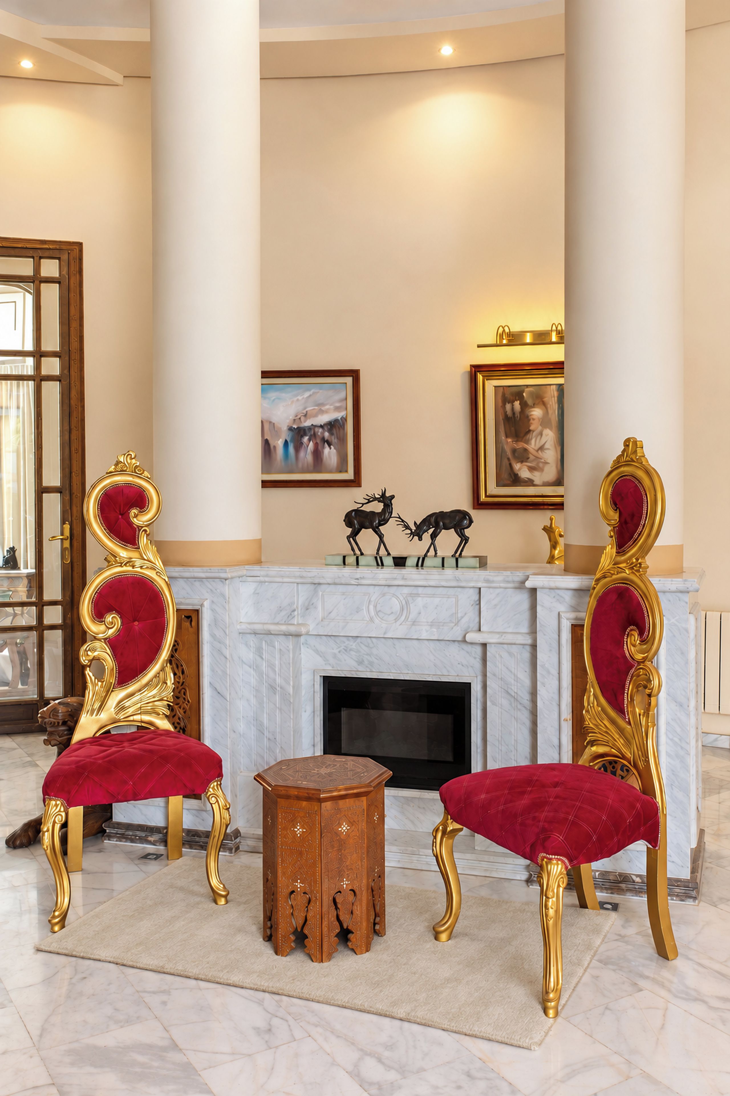A luxurious room with a marble fireplace, two ornate golden chairs with red velvet upholstery, a small wooden side table, and a framed painting above the fireplace.