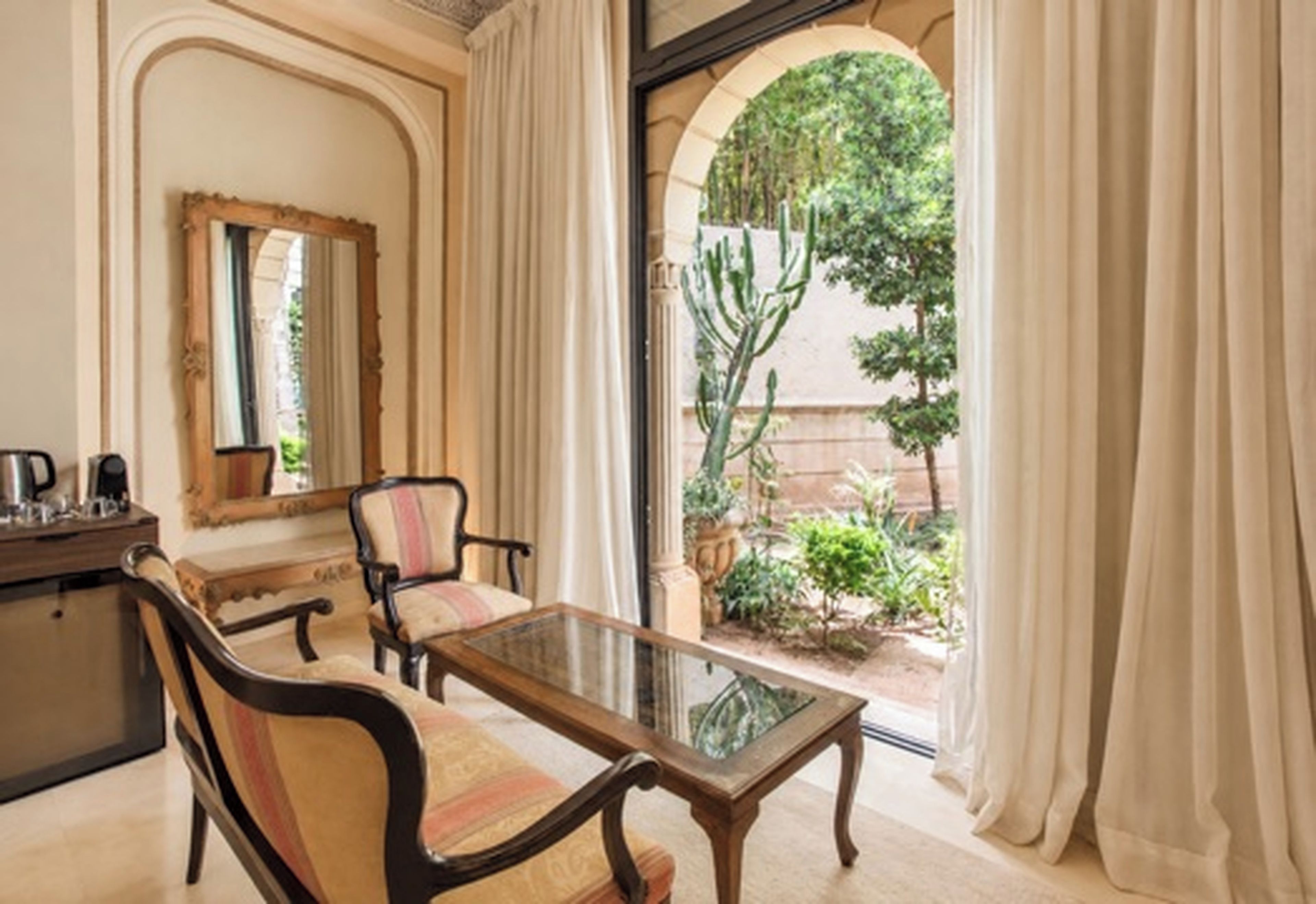 A room with a glass-top table, two chairs, a small desk with a mirror, and a view of a garden through a doorway.