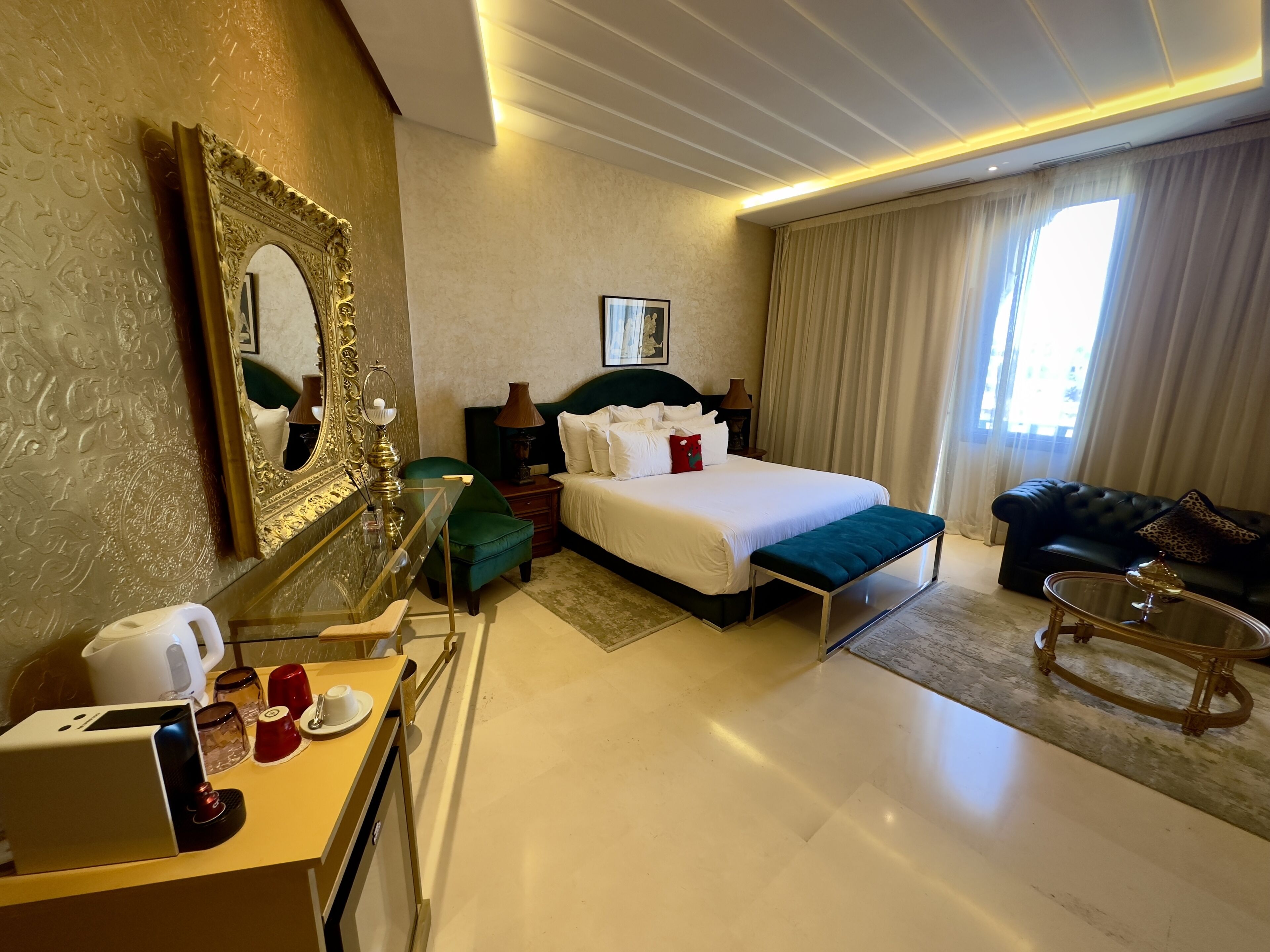 A hotel room with a large bed, a green armchair, a coffee table, a sofa, and a desk with a coffee maker and cups.