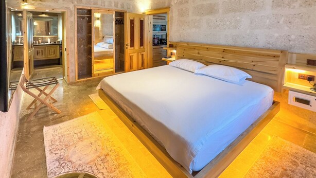 Un dormitorio con una cama grande, cabecera de madera, mesitas de noche y un espejo. Hay una silla de madera y una pequeña mesa con una lámpara. La habitación tiene una pared de piedra y un techo de madera.