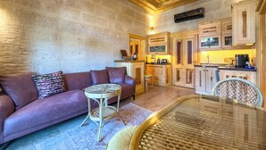 Una sala de estar acogedora con un sofá, una mesa de centro de vidrio y un sillón de mimbre. Se observa un área de cocina con armarios de madera y una lámpara colgante.