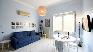 A living room with a blue sofa, a small round table with chairs, a white radiator, and a window with natural light. - STANDARD APARTMENT (Roma)