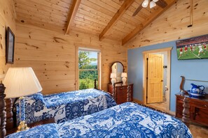 A room with wooden walls and ceiling, two beds with blue floral bedding, a wooden dresser, a mirror, and a door leading outside.