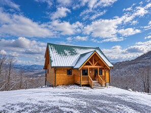 A wooden cabin with a green roof, surrounded by snow-covered landscape and mountains.