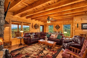 A cozy cabin interior with a leather sofa, a fireplace, and a large window offering a view of the outdoors.