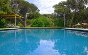 Relax by the serene pool surrounded by lush greenery and shaded by a pergola.