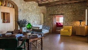 A rustic room with a stone wall, a wooden table with books and a green vase, a yellow armchair, and a red sofa.