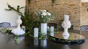 A dining table with white floral centerpieces, surrounded by chairs with black lattice backs, set against a stone wall with greenery.