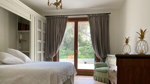 A bedroom with a large bed, a wooden dresser, and a green chair. There is a window with curtains, a bookshelf, and a chandelier.