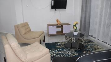 Una sala de estar con un sillón beige, un sofá de cuero negro, una mesita redonda con un jarrón de flores, un aparador de madera y un televisor de pantalla plana montado en la pared.