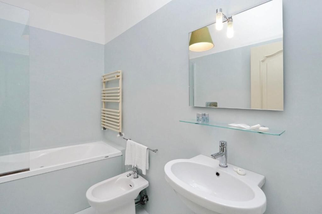 A modern bathroom with a bathtub, two sinks, a towel rack, a glass shelf with toiletries, and a mirror with a light fixture.