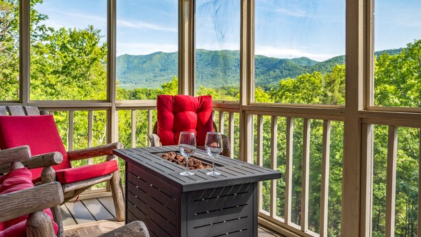 Eine Holzterrasse mit einem roten Stuhl und einem Tisch mit zwei Weingläsern, mit Blick auf eine gebirgige Landschaft.