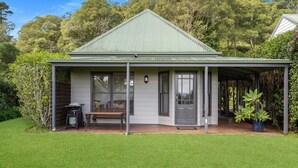 A single-story house with a green roof, a covered porch, and a small garden area with a bench and a grill. - Watta Cottage - Kangaroo Valley (Wattamolla)