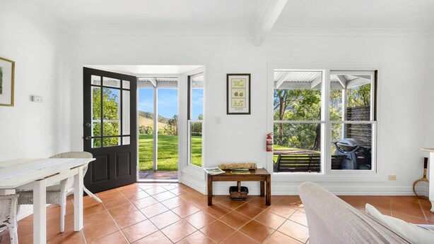A spacious room with a large window offering a view of a green landscape, a door leading outside, and a small table with a basket. - Watta Cottage - Kangaroo Valley (Wattamolla)