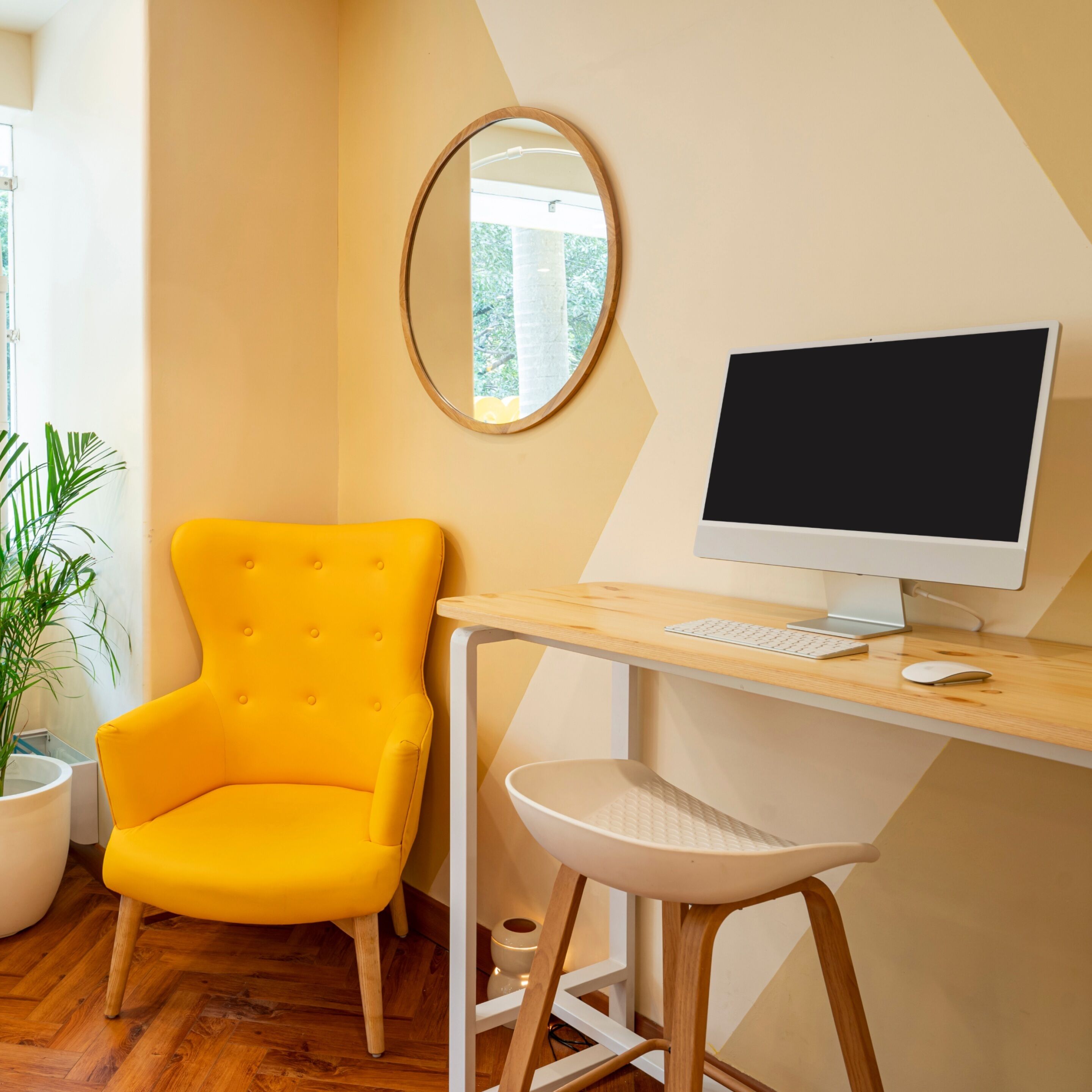 Ein Zimmer mit einem Holztisch, einem gelben Stuhl, einem weißen Hocker, einem runden Spiegel und einem Computermonitor.