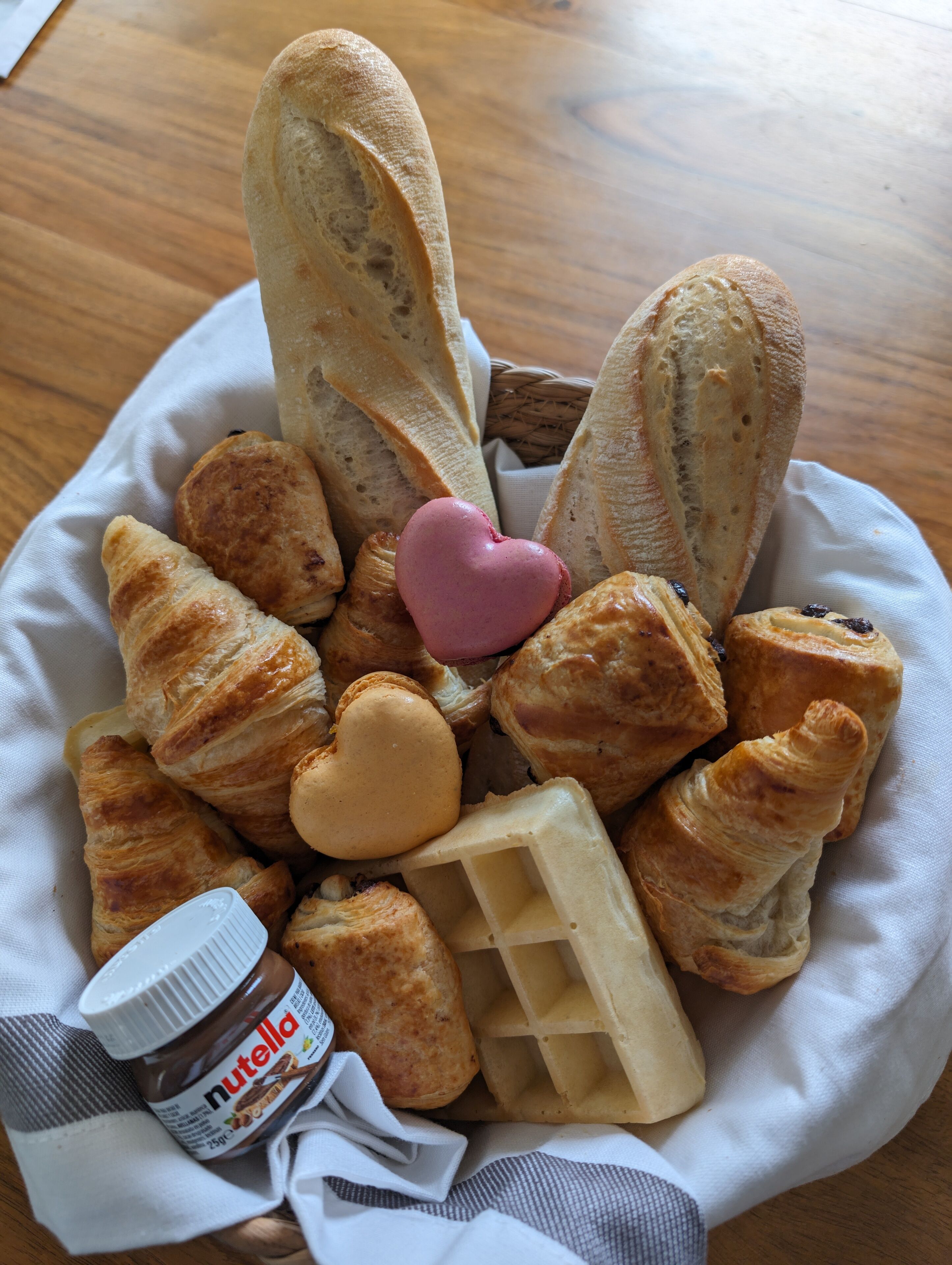 Een mandje met diverse gebakjes, waaronder croissants, een hartvormige koek en een wafel, met een pot Nutella ernaast.