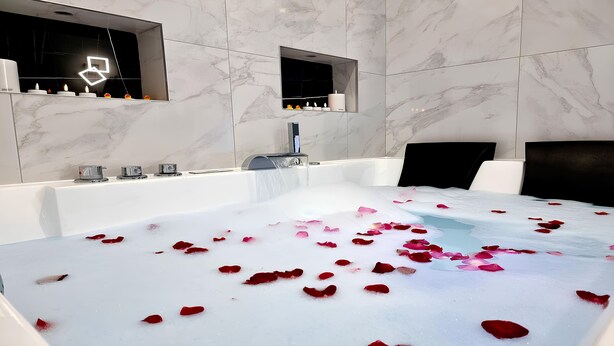 Un baño moderno con una bañera grande llena de agua y pétalos de rosa, un espejo y un estante en la pared con velas.
