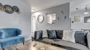Un salon moderne avec un fauteuil bleu, un canapé gris agrémenté de plusieurs coussins, une table basse en bois et des œuvres d’art décoratives accrochées au mur.
