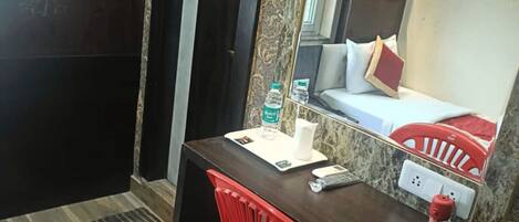 A compact hotel room with a dark wooden desk, a red chair, a small table with a water bottle, a cup, and a remote control. A large mirror reflects a bed with red and white bedding.