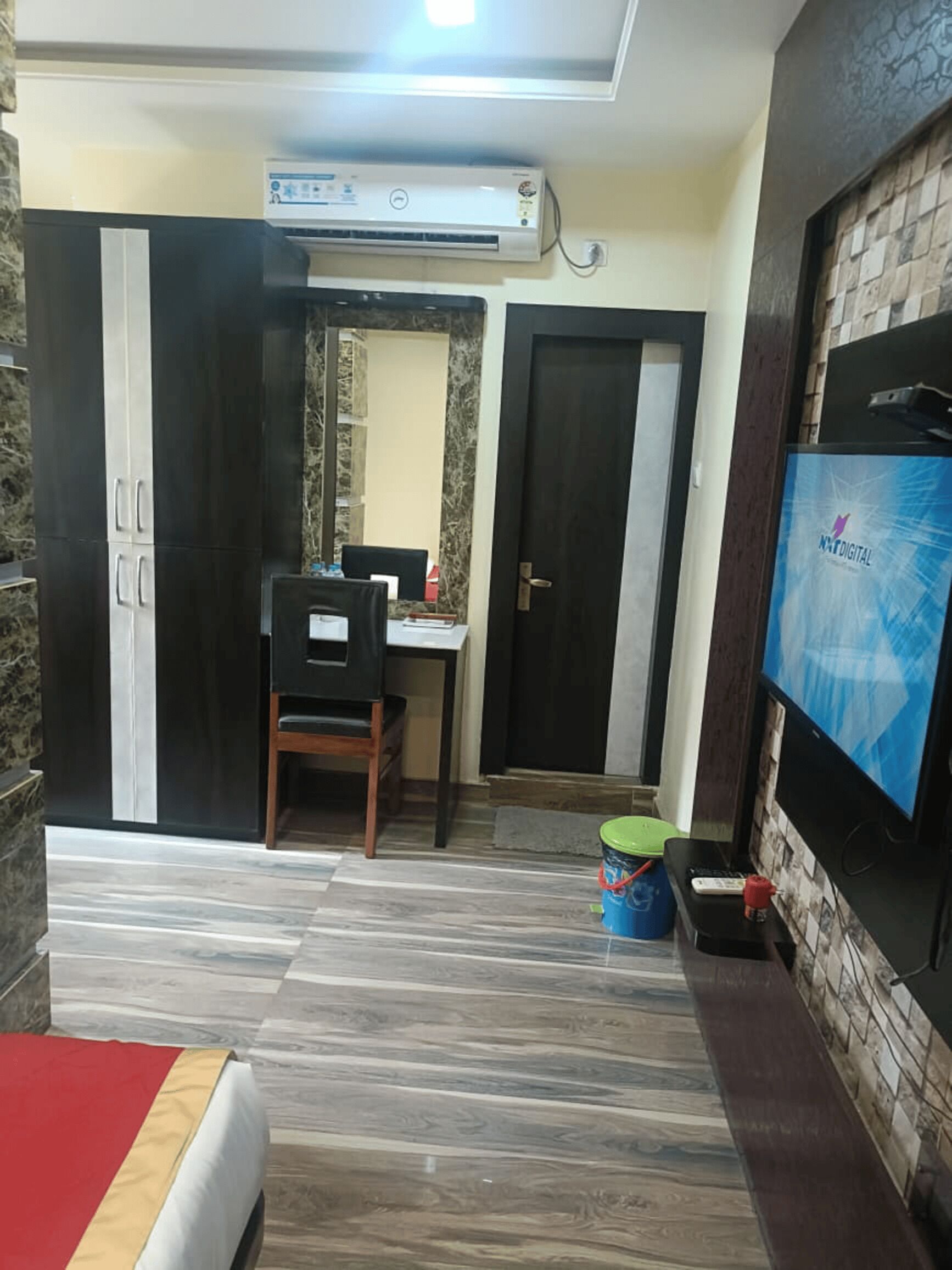 A hotel room with a bed, a desk with a chair, an air conditioning unit, a television, and a small table with a bucket.