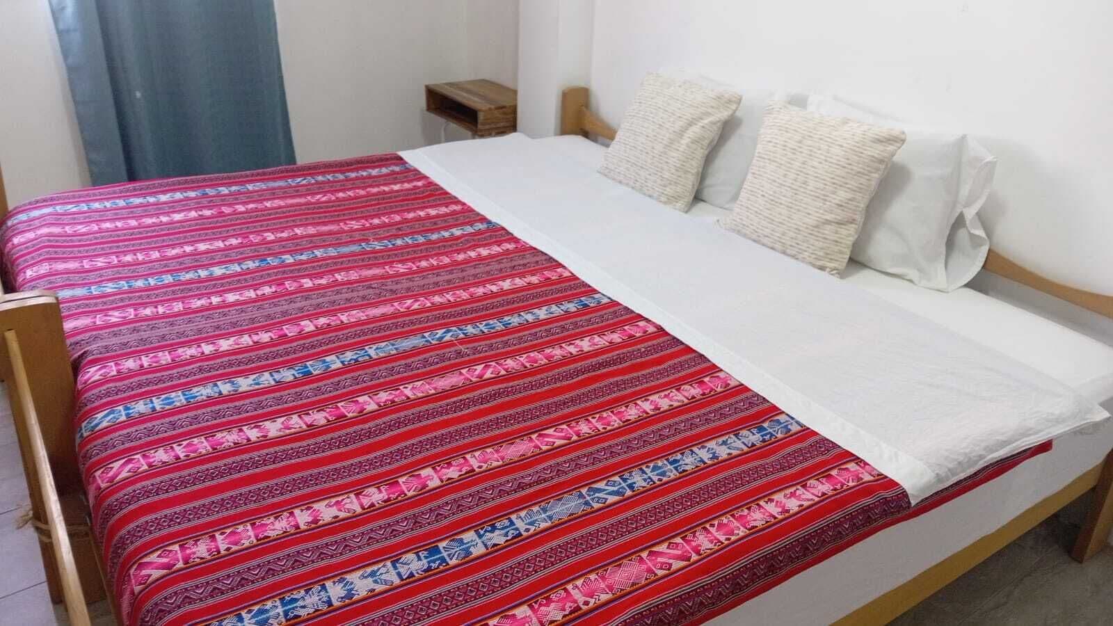 Una cama con colcha y almohadas estampadas, cabecera de madera y una mesita de noche pequeña de madera.