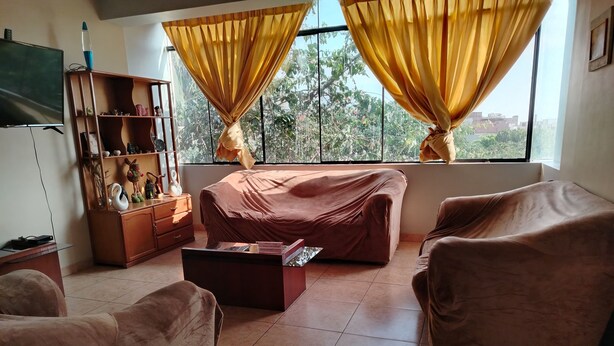 A living room with a sofa, a coffee table, and a TV. There are curtains on the windows, and a bookshelf with decorative items. - Chakana Family Home (Callao)